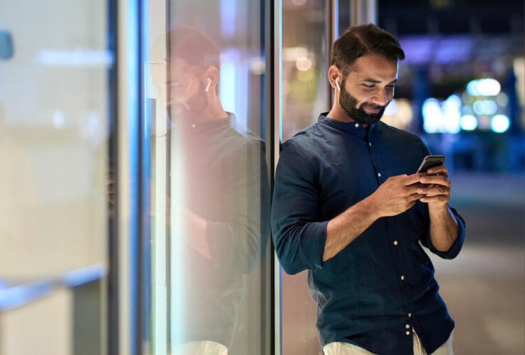 man leaning against window with earpods in and smiling at his phone at night